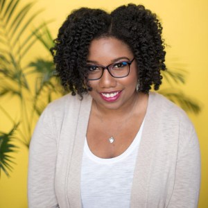 Photo of the author of SLAY, Brittney Morris. She has natural hair, done in a twist out, black glasses, and pinkish red lipstick. She's wearing a white T-shirt with a cream cardigan over it. 